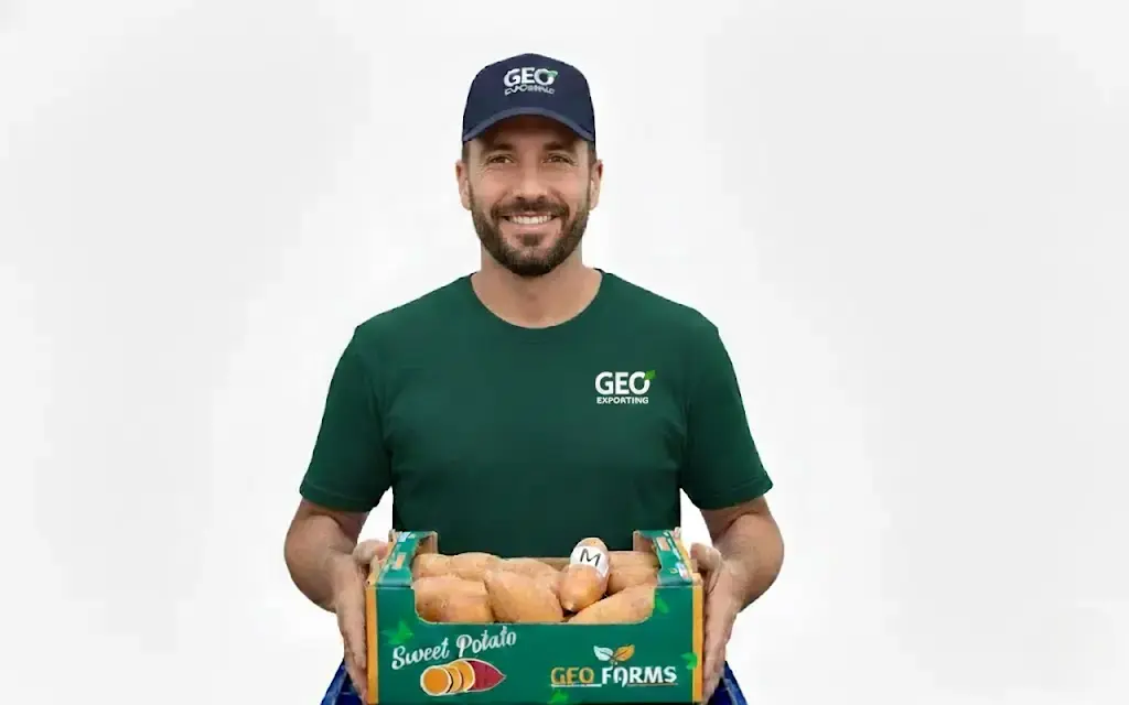 A smiling GEO Exporting staff member holding a 6kg carton of premium Egyptian sweet potatoes, featuring the GEO Farms brand logo and medium-sized (M) calibration.